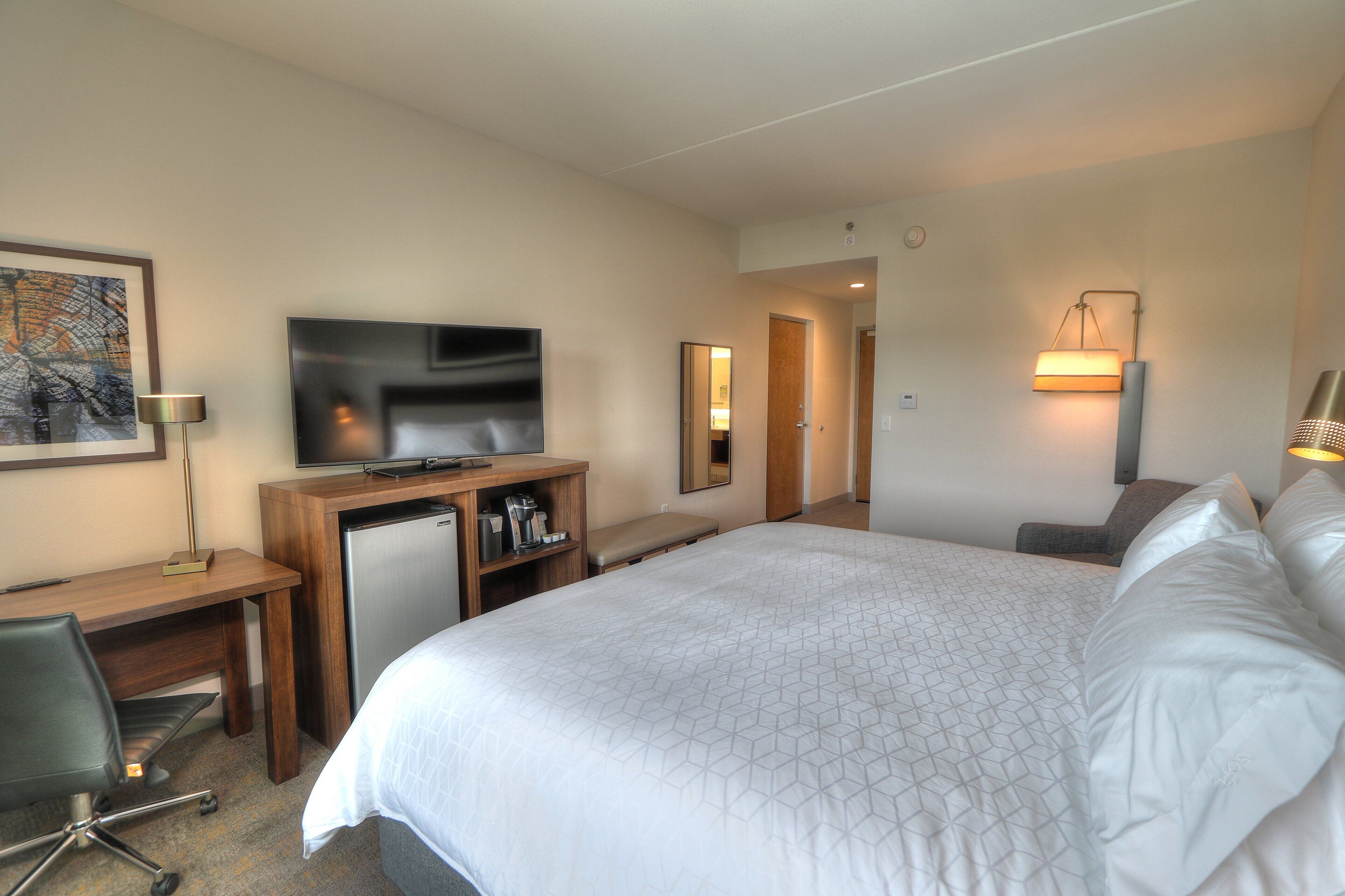 A hotel room with a bed, a desk with a television, a coffee machine, and a chair.