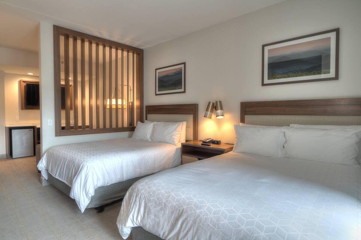 A hotel room with two beds, a wooden partition, a desk with a lamp, and a small fridge.