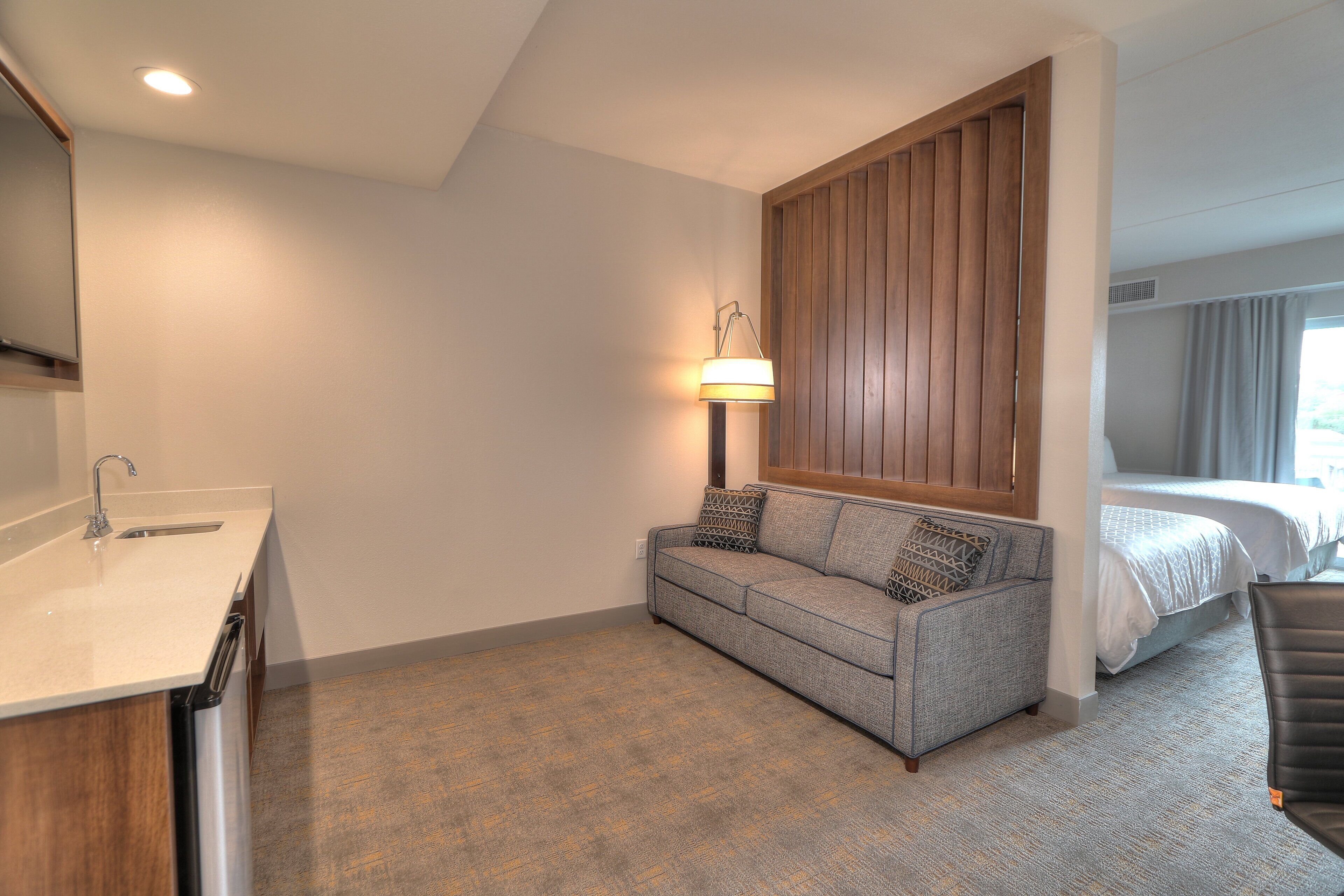 A hotel room with a kitchenette, a grey sofa with patterned cushions, a wooden panel wall, and a bed with a white duvet.