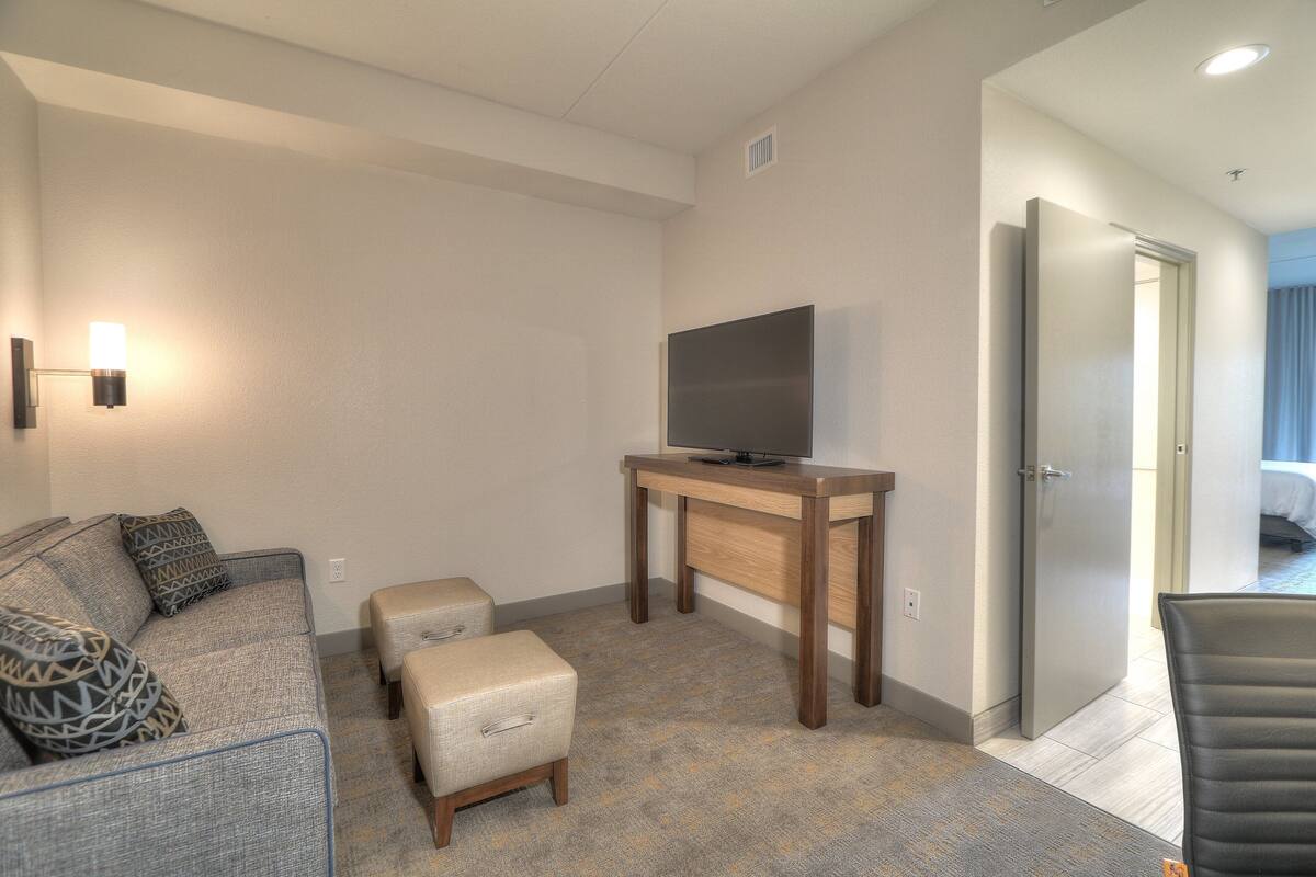 A hotel room with a gray sofa, a small ottoman, a wooden table with a flat-screen TV, and a door leading to a bedroom.