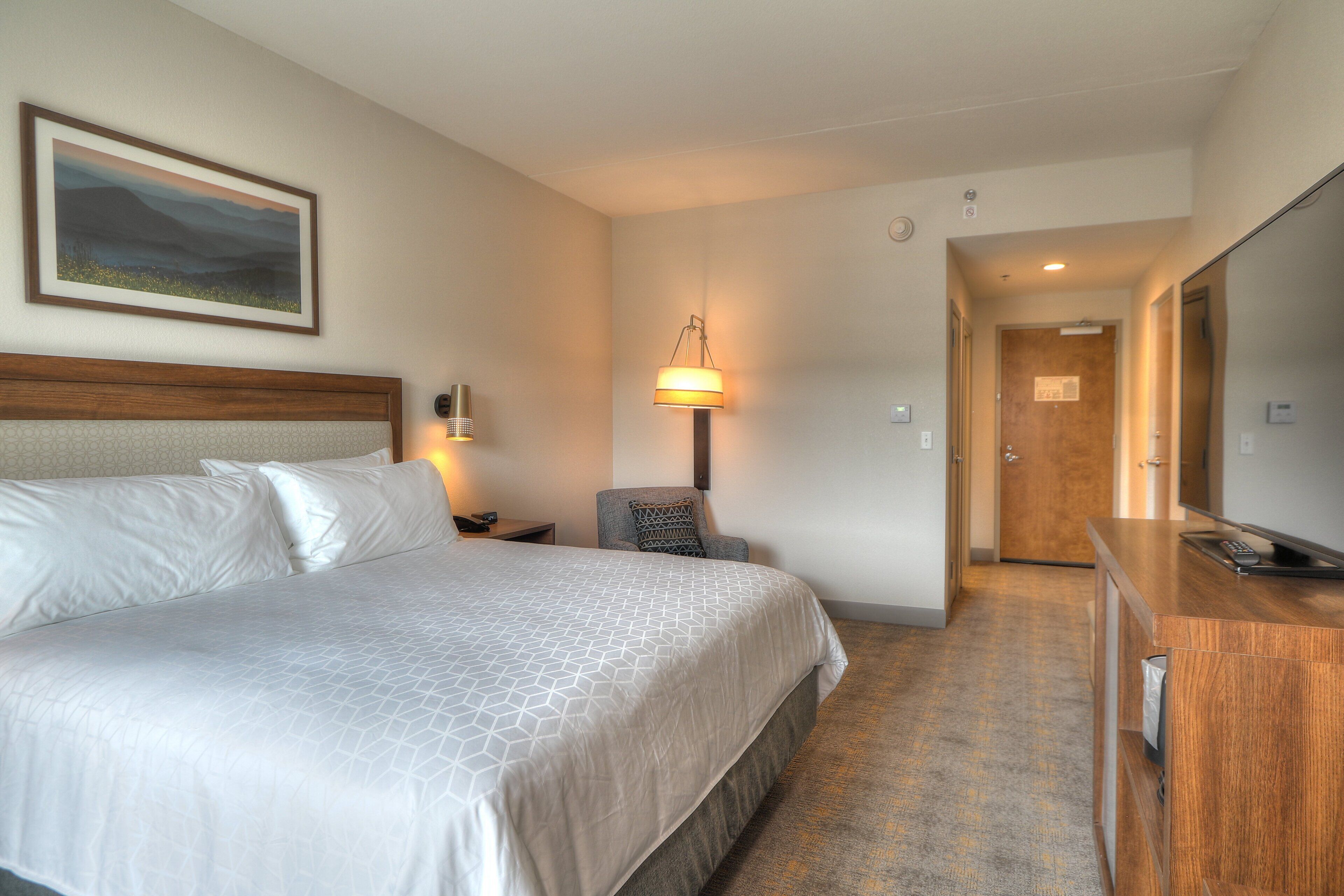 Habitación de hotel con una cama grande, lámparas de noche, un escritorio con control remoto y una puerta que da a otra habitación.