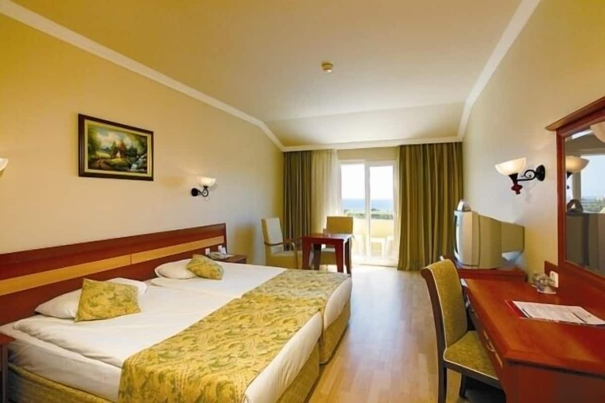 A hotel room with two beds, a desk, and a chair. There is a painting on the wall, a window with curtains, and a view of the outside.
