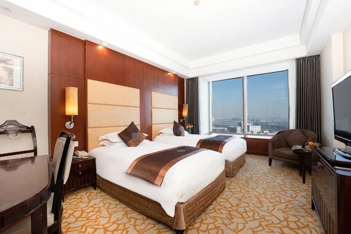 A hotel room with two beds, a desk, a chair, a television, and a large window with a city view.