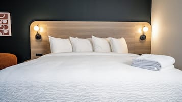 Een netjes opgemaakt bed met wit beddengoed en kussens, twee wandlampen voor leeslicht en een opgevouwen witte handdoek aan de zijkant.