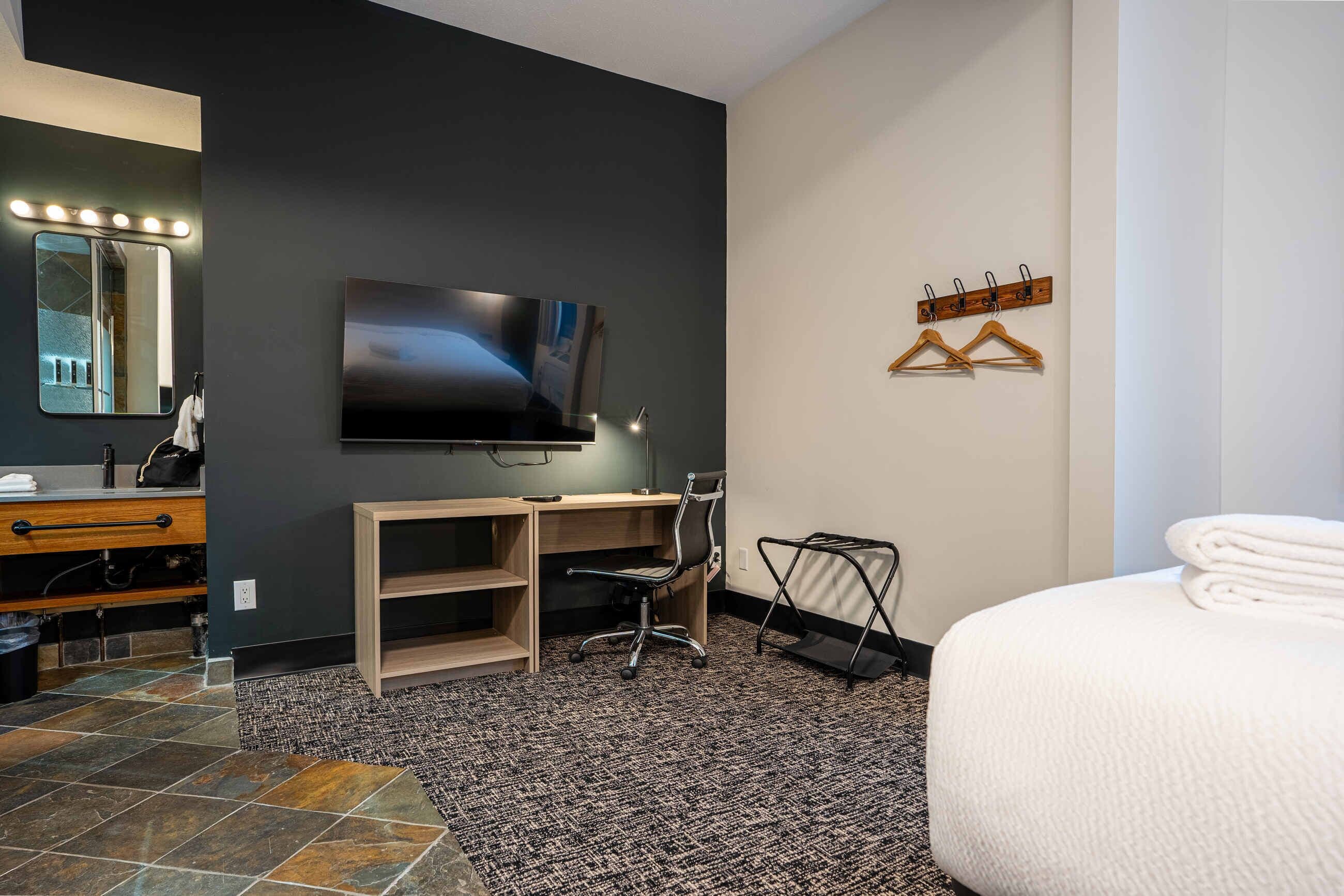 A modern hotel room with a large flat-screen TV, a desk with a chair, a bed with a neatly folded white towel, and a mirror with lights.