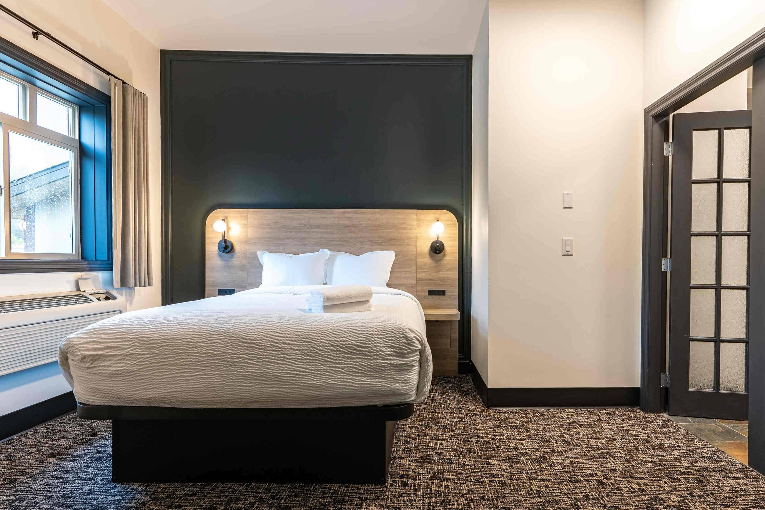 Habitación de hotel con una cama grande, cabecera oscura, dos luces fijadas a la pared, una ventana con cortinas y una puerta con paneles de vidrio esmerilado.