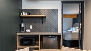 Una kitchenette compacta con microondas, cafetera y un refrigerador pequeño. Hay un estante de madera sobre el microondas y se ve una cama a través de una puerta abierta.