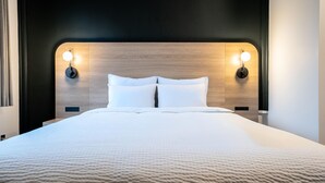 Una cama bien hecha con cabecera de madera, dos lámparas de lectura fijadas en la pared y un enchufe en la cabecera.