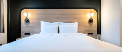 Uma cama bem arrumada com cabeceira de madeira, duas luminárias de leitura fixadas na parede e uma tomada na cabeceira.