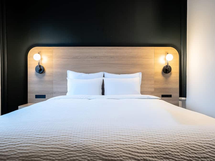 Ein ordentlich bezogenes Bett mit Holz-Kopfteil, zwei Wandleuchten zum Lesen und einer Steckdose am Kopfteil.