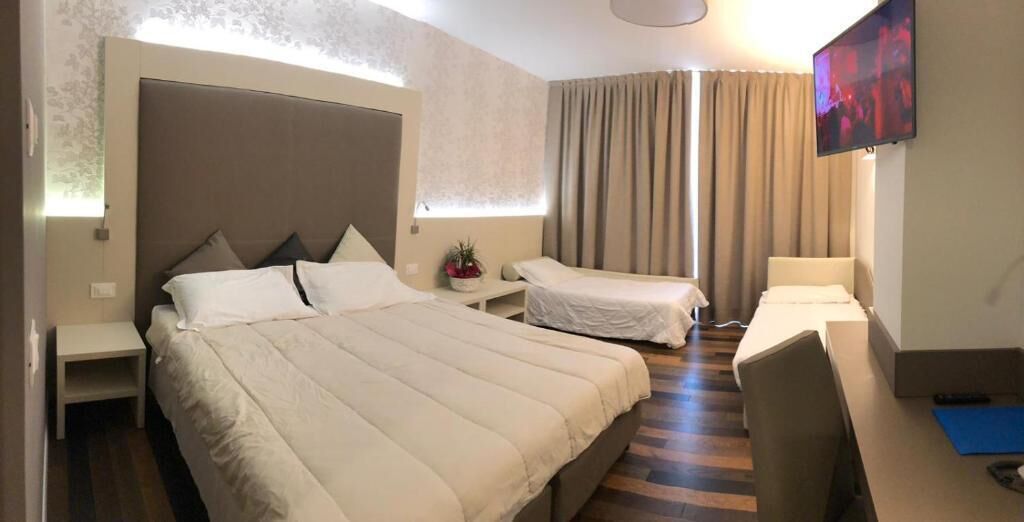 A hotel room with a large bed, a smaller bed, a desk, a chair, a TV, and a window with curtains.