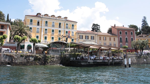 A waterfront building with multiple balconies, a covered outdoor seating area, and a stone wall along the water. - Hotel Excelsior Splendide (Bellagio)