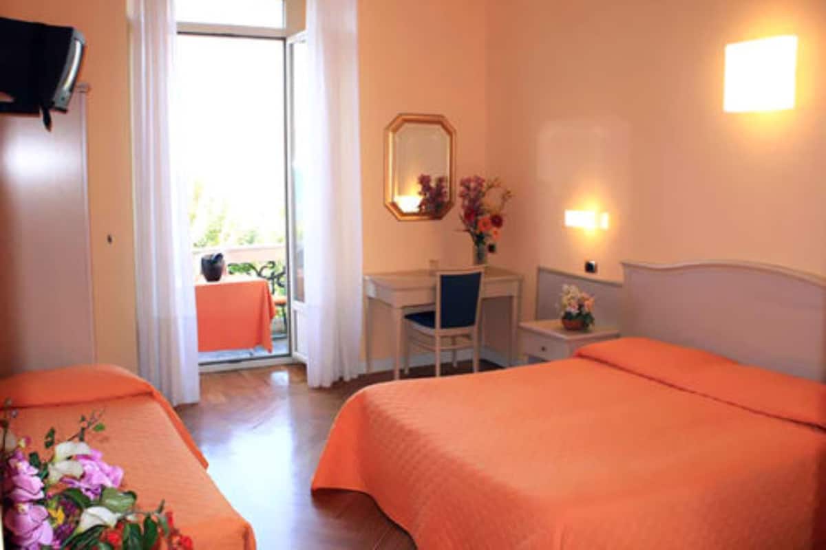 A hotel room with two beds, a desk, a chair, a small table with a vase of flowers, and a window with curtains.