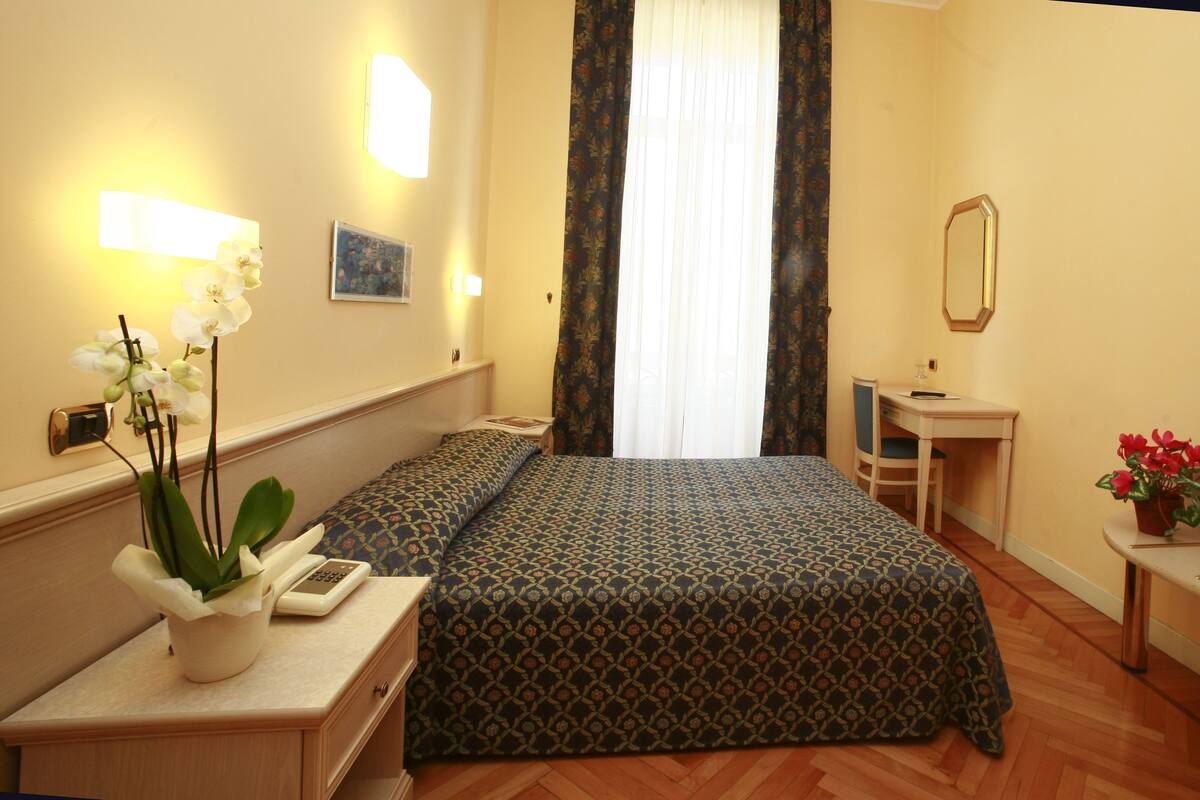 A hotel room with a bed, bedside table, lamp, and a small desk with a chair.