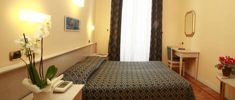 A hotel room with a bed, bedside table, lamp, and a small desk with a chair.