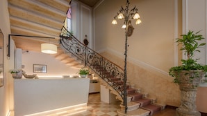 A hotel lobby with a checkered floor, a reception desk, a staircase with ornate railings, and a large potted plant. - Hotel Excelsior Splendide (Bellagio)