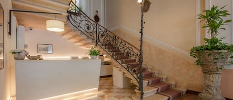 Un vestíbulo de hotel con suelo a cuadros, una mesa de recepción, una escalera con barandilla ornamentada y una planta grande en maceta.