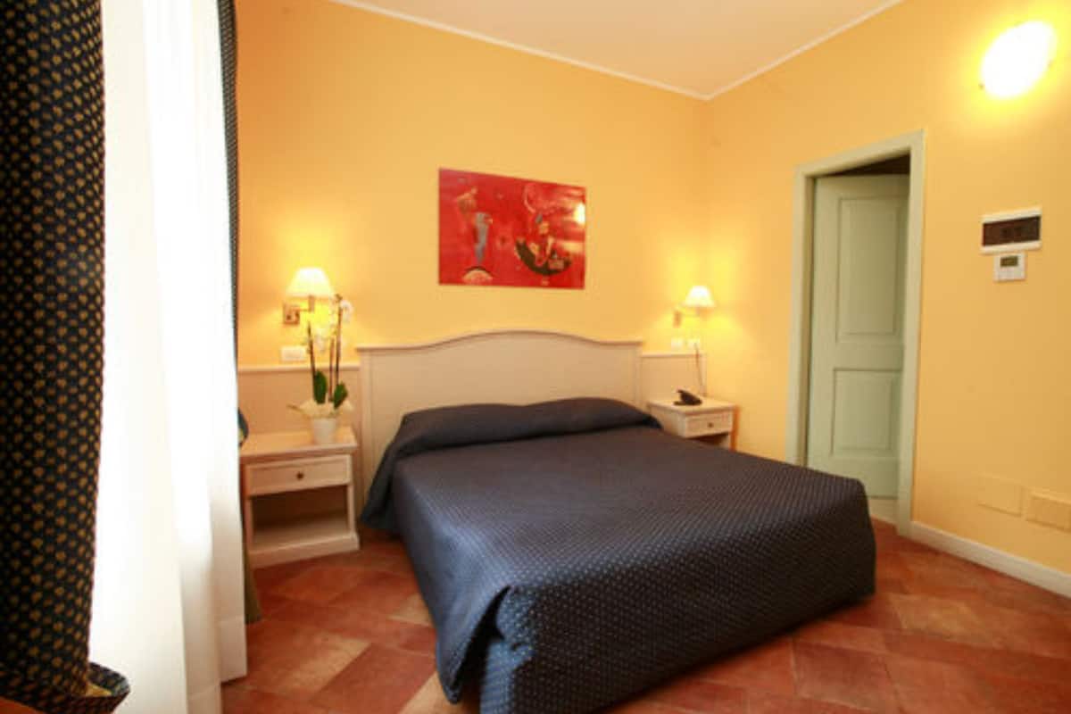 A hotel room with a bed, two bedside tables with lamps, a red painting on the wall, and a green door.