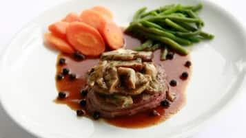 A plate with a steak topped with mushrooms and sauce, accompanied by sliced carrots and green beans.