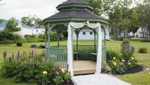 A gazebo with a green roof and white drapery, surrounded by a garden with various flowers and a bench. - LimeRock Inn (Rockland)
