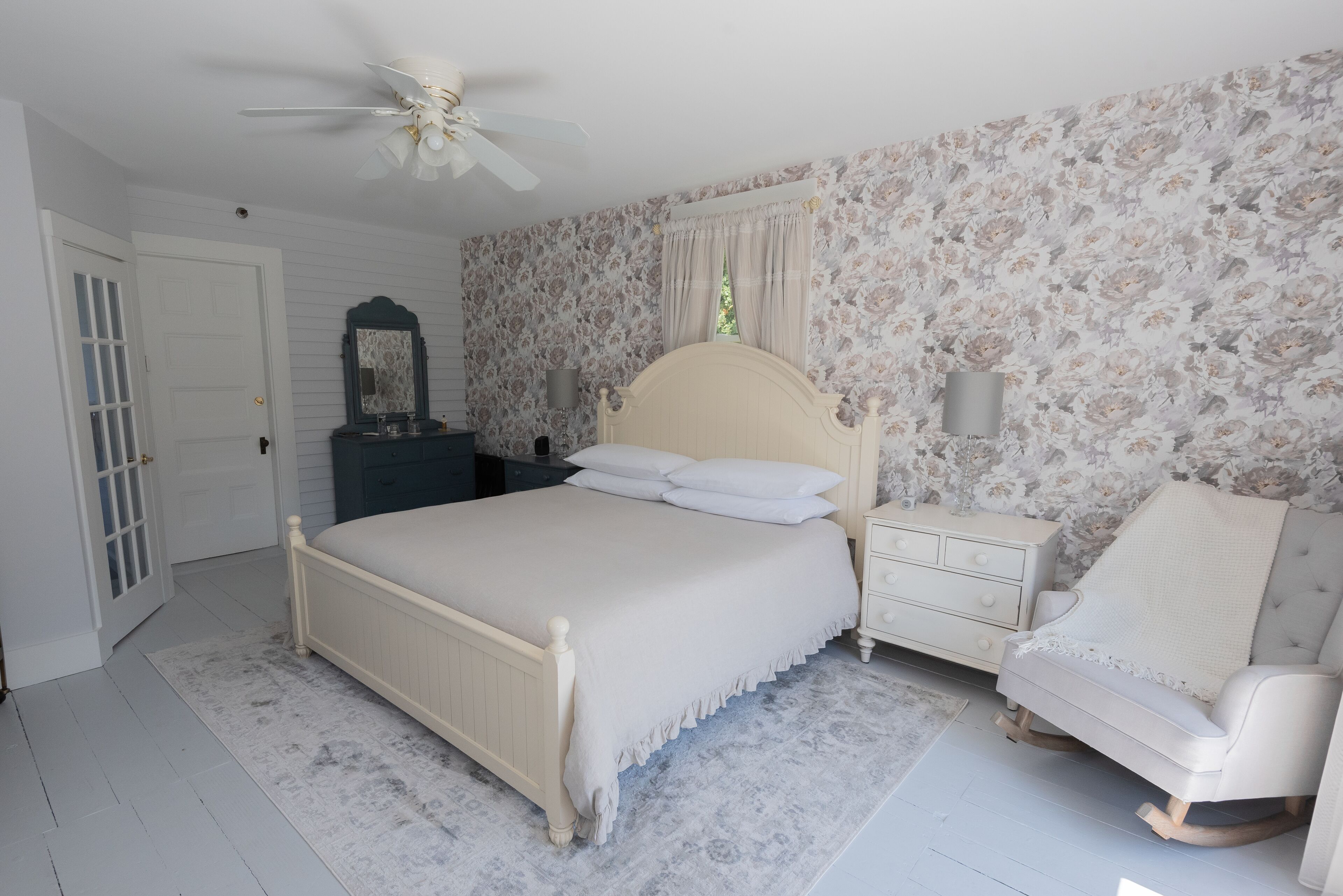 A bedroom with a bed, nightstands, a chair, a ceiling fan, and floral wallpaper.