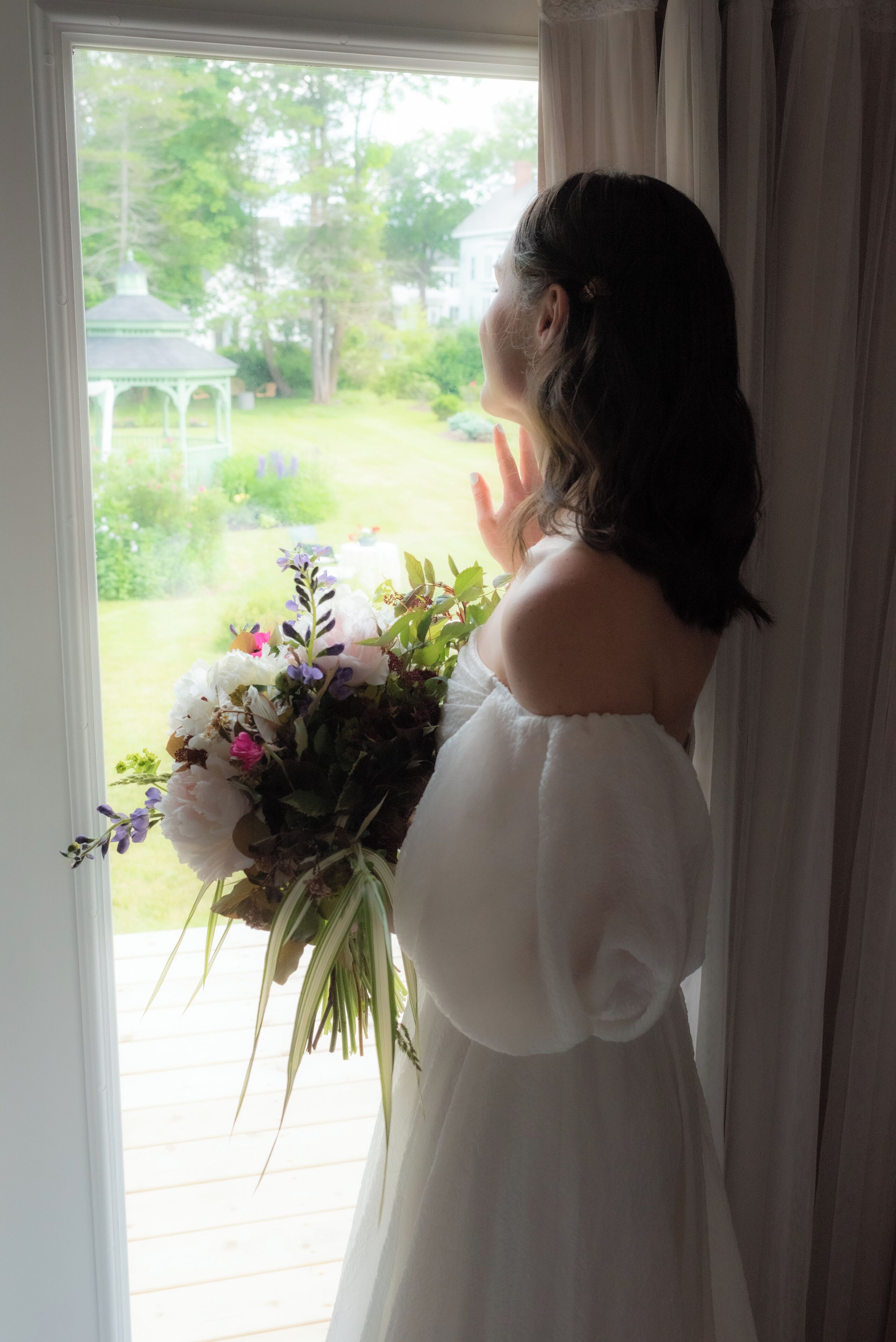A person holding a bouquet of flowers, standing by a window with a view of a gazebo and greenery.