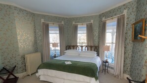A bedroom with a large bed, two windows with curtains, a radiator, a small table with a lamp, and a chair. - LimeRock Inn (Rockland)
