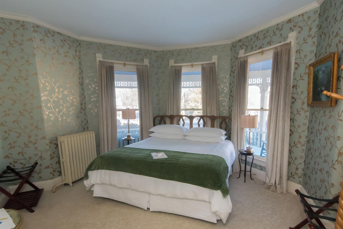 Un dormitorio con una cama grande, dos ventanas con cortinas, un radiador, una mesita con lámpara y una silla.