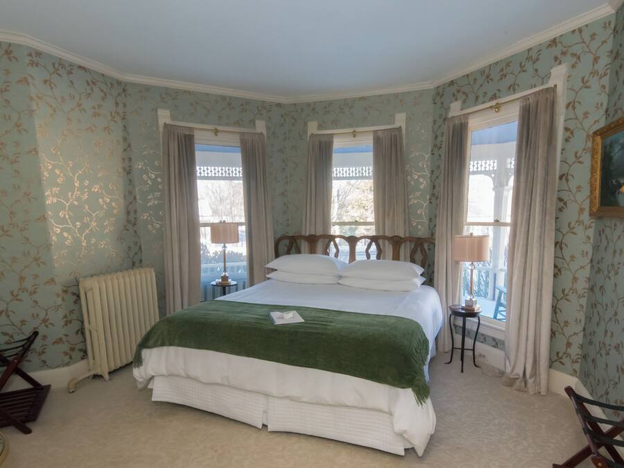 Une chambre avec un grand lit, deux fenêtres avec des rideaux, un radiateur, une petite table avec une lampe et une chaise.