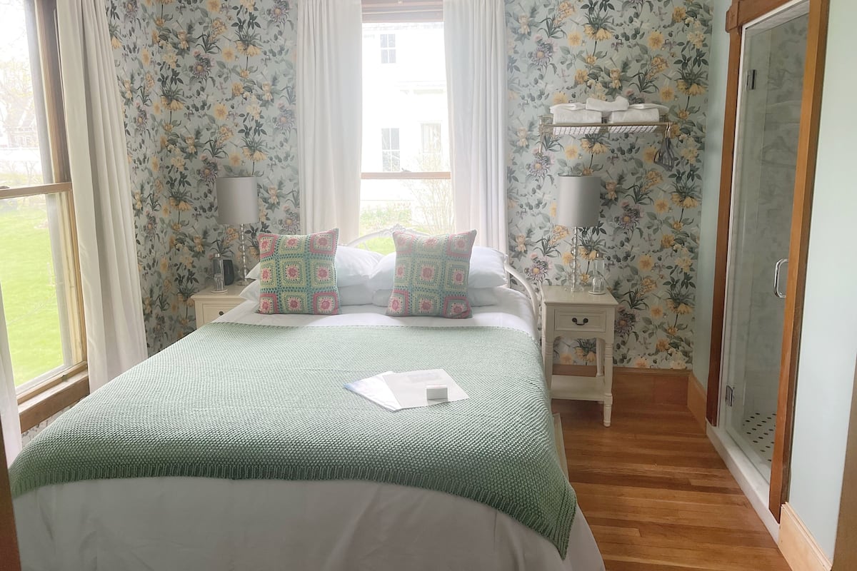 Un dormitorio con papel tapiz floral, una cama con cubrecama verde, dos almohadas decorativas, una mesita de noche con lámpara y una ventana con cortinas.