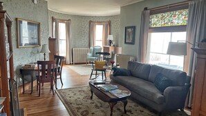 A living room with a sofa, chairs, a coffee table, and a radiator. There are windows with curtains and a framed picture on the wall. - LimeRock Inn (Rockland)