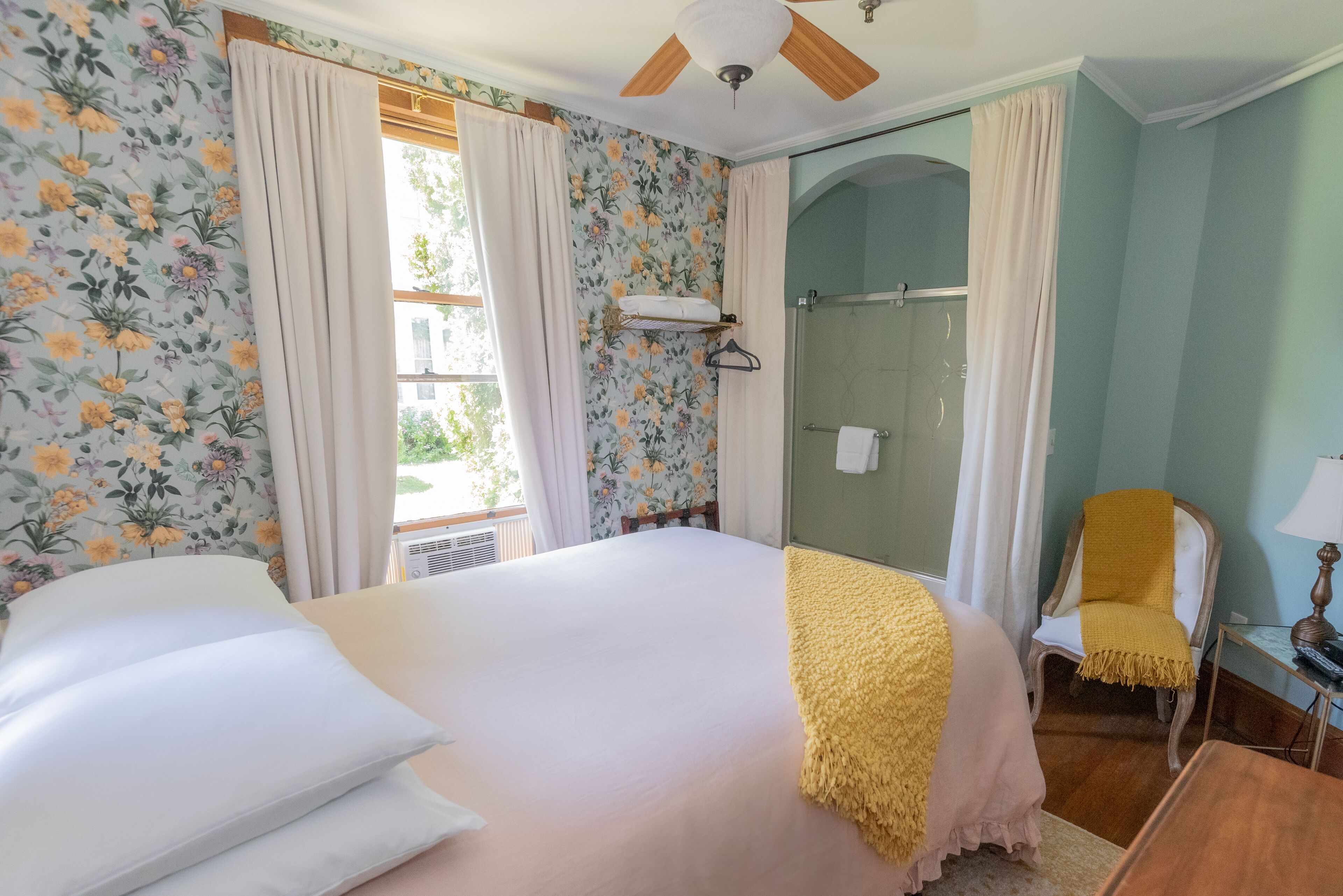 A bedroom with a bed, a chair, a small table, a ceiling fan, and a window with curtains.