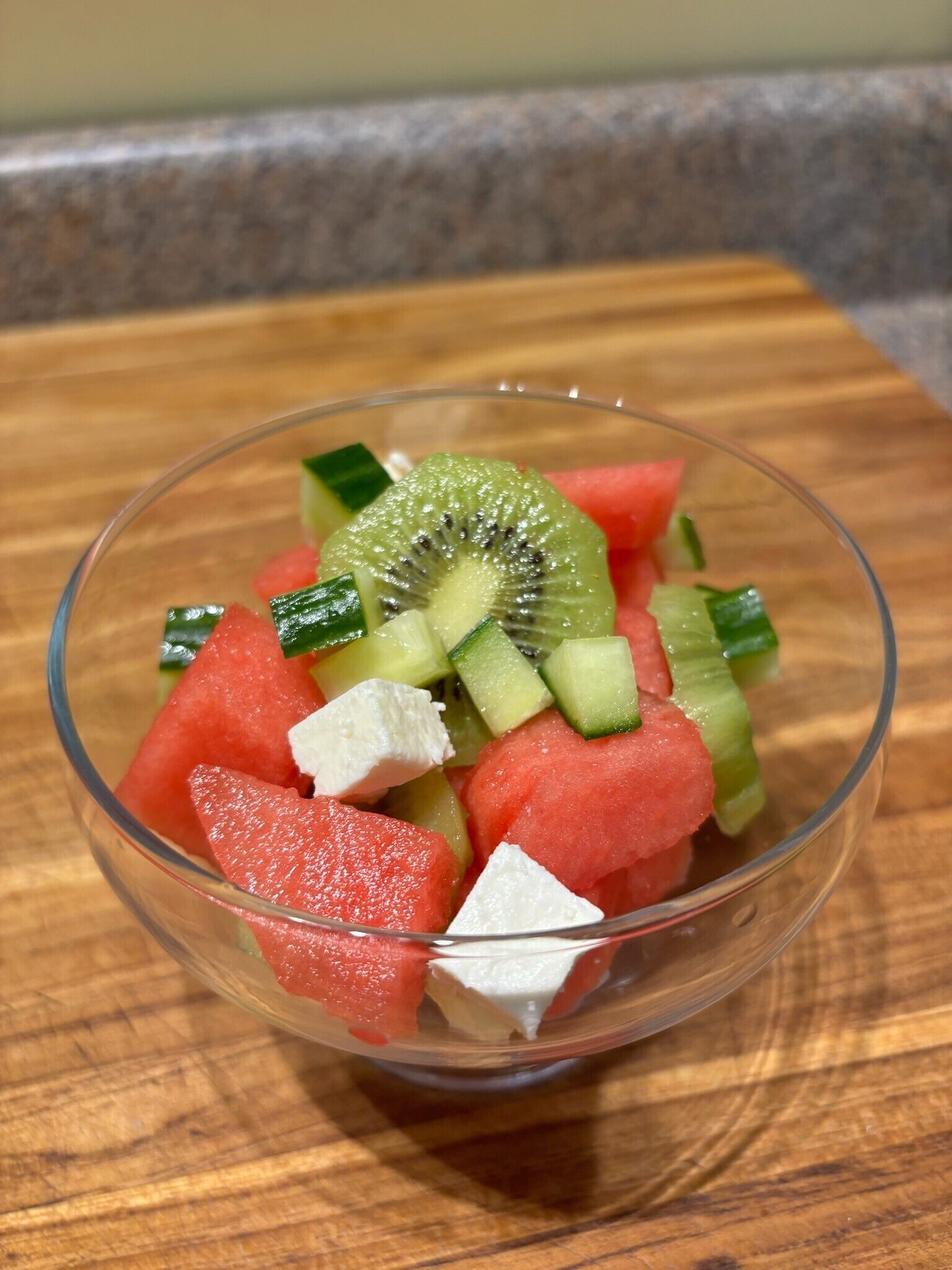 A bowl of fruit salad with watermelon, kiwi, cucumber, and cheese on a wooden cutting board.