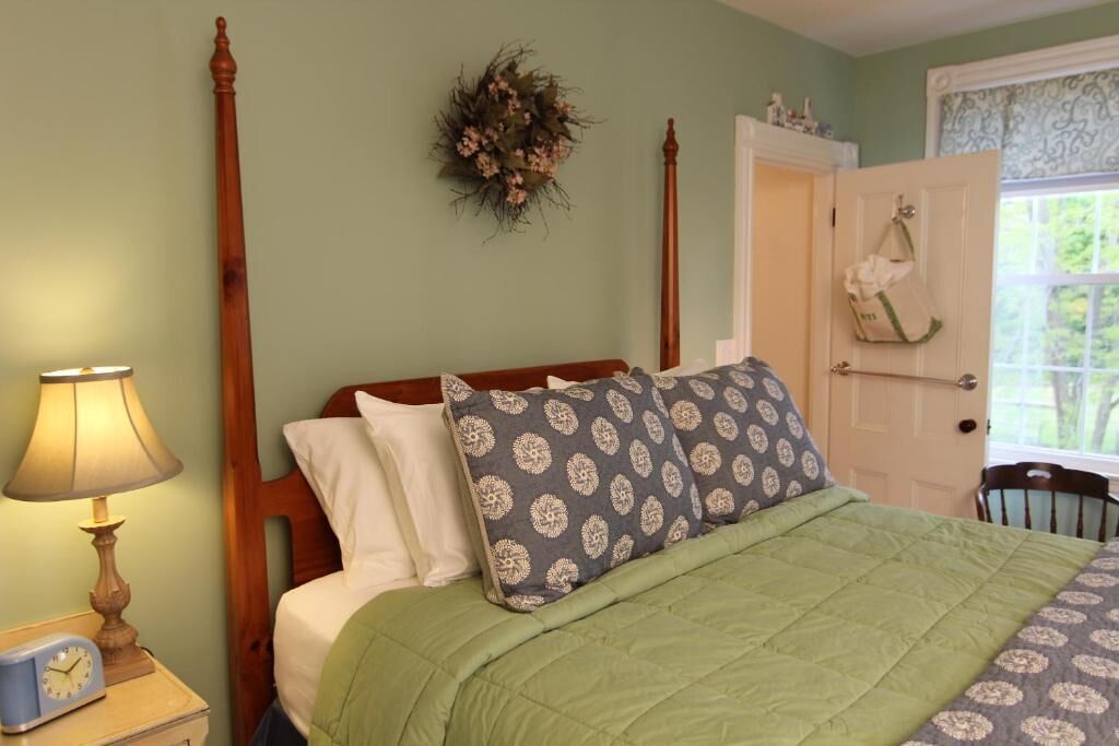 A bedroom with a wooden four-poster bed, green bedding, a nightstand with a lamp, and a wall-mounted floral decoration.