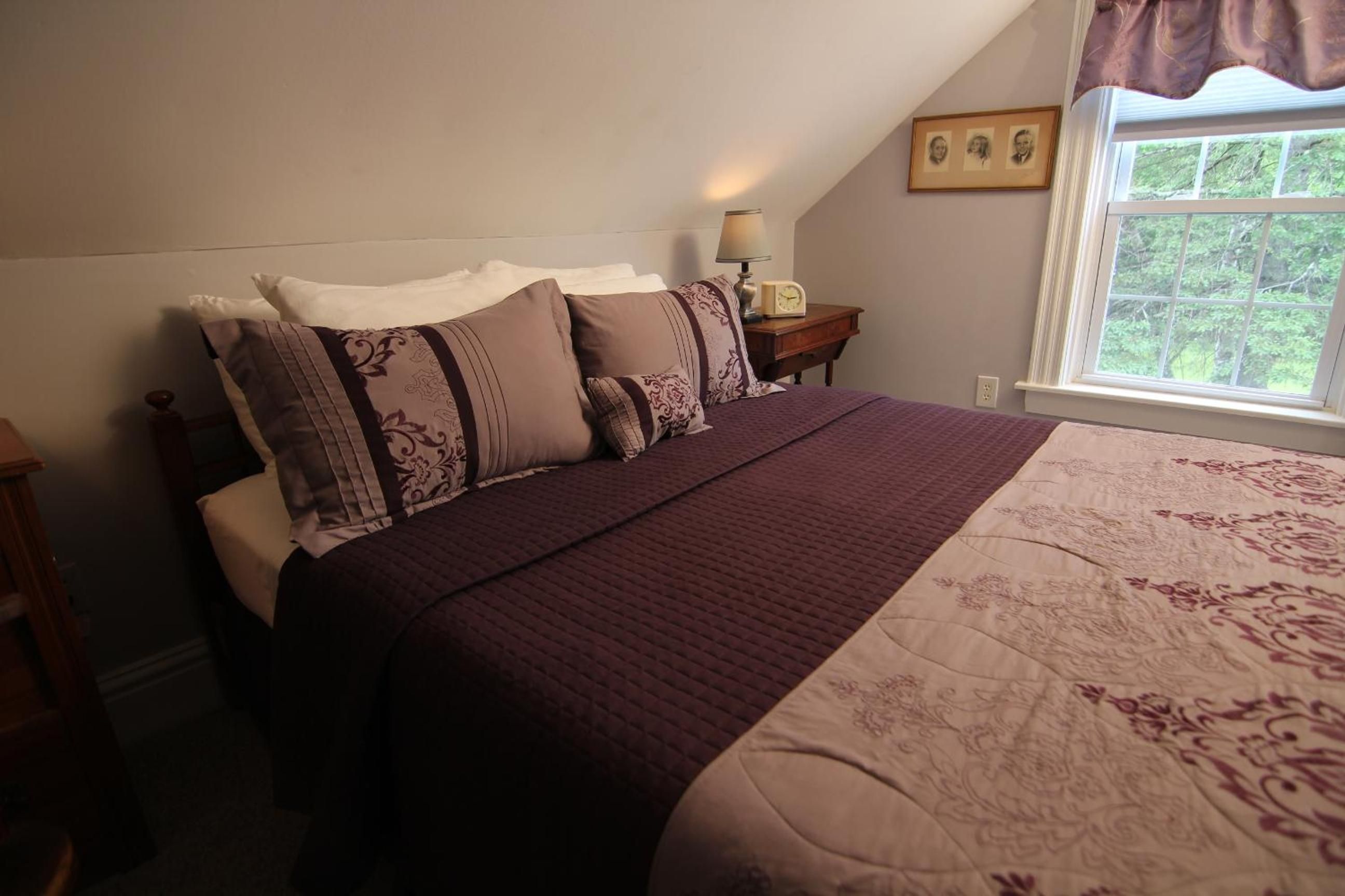 A bedroom with a large bed, a nightstand with a lamp, and a window with a view of greenery.