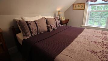 A bedroom with a large bed, a nightstand with a lamp, and a window with a view of greenery.
