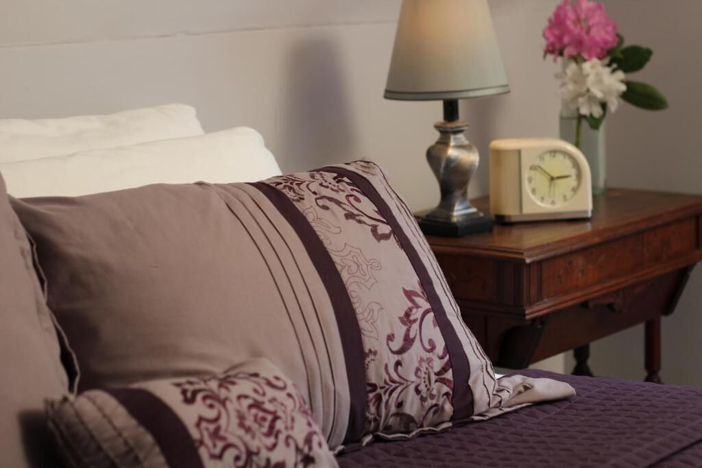 A bed with patterned pillows, a bedside table with a lamp, a clock, and a vase with flowers.