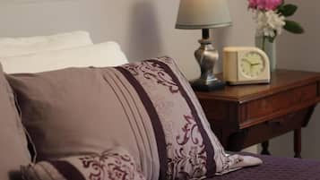 A bed with patterned pillows, a bedside table with a lamp, a clock, and a vase with flowers.