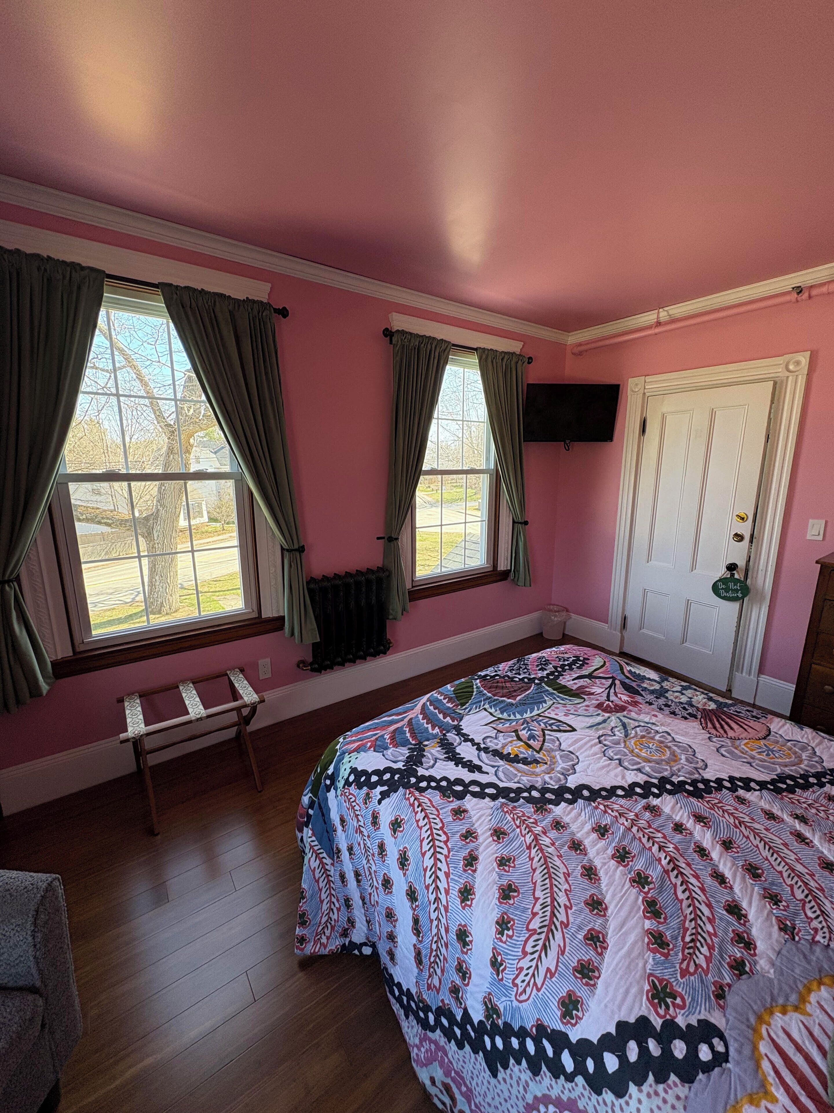 Ett sovrum med rosa väggar, två fönster med gardiner, en säng med ett mönstrat täcke, en bänk och en dörr med en skylt.
