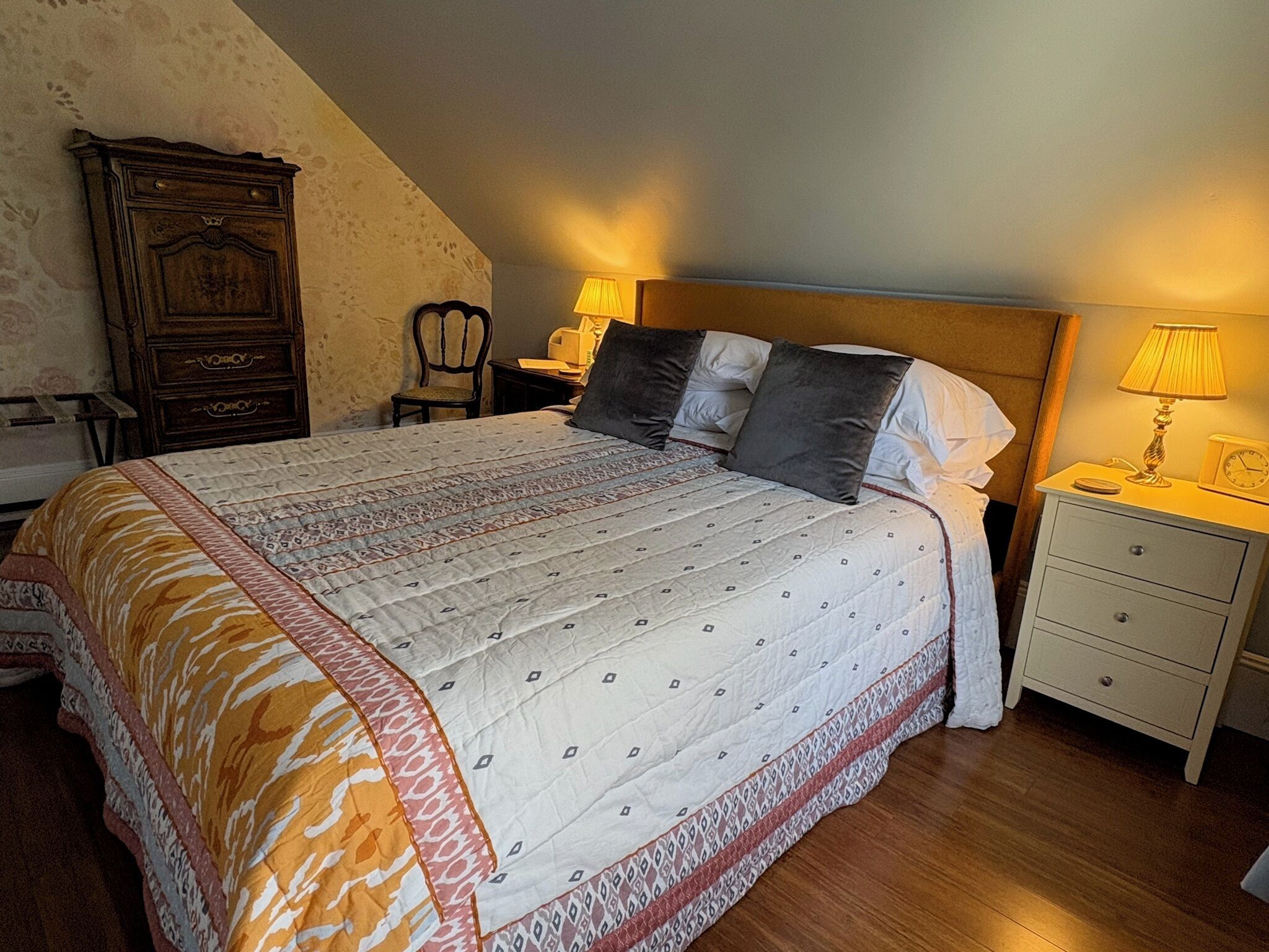 A bedroom with a large bed, a wooden dresser, a chair, and a nightstand with a lamp.