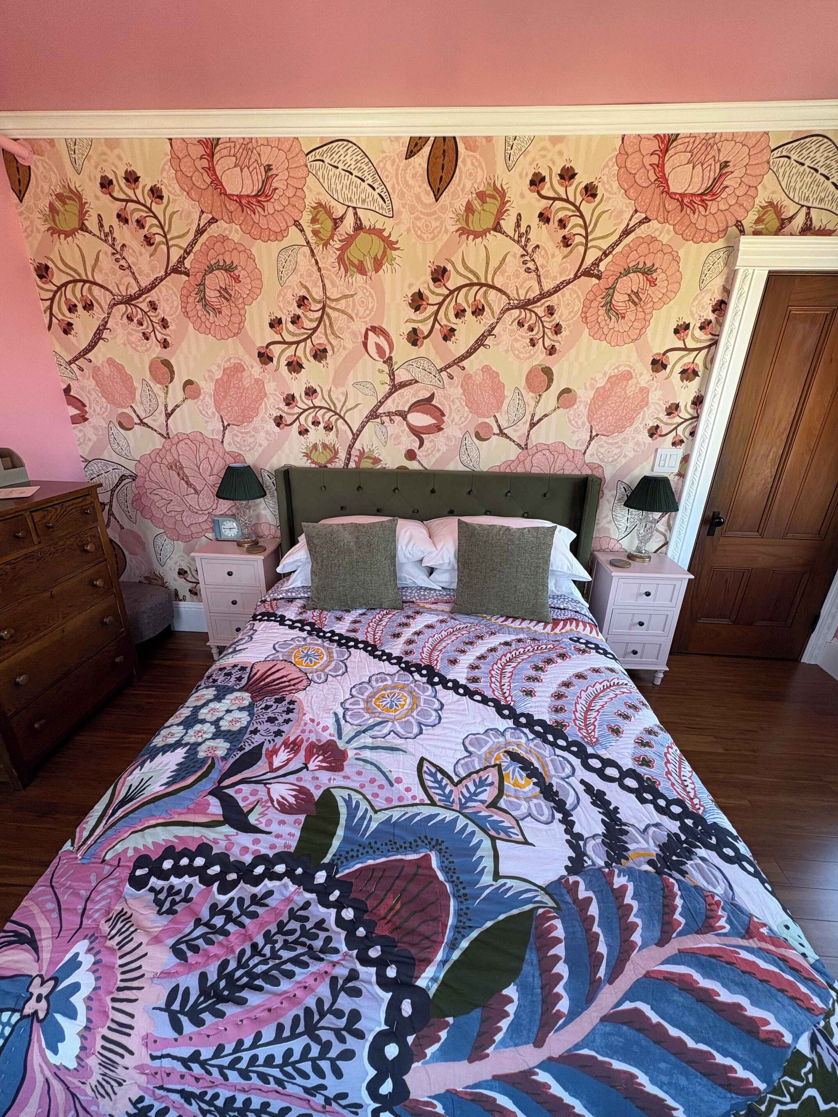 Un dormitorio con pared de estampado floral, cabecera tapizada en verde, colcha estampada, dos mesitas de noche con lámparas y un aparador de madera.