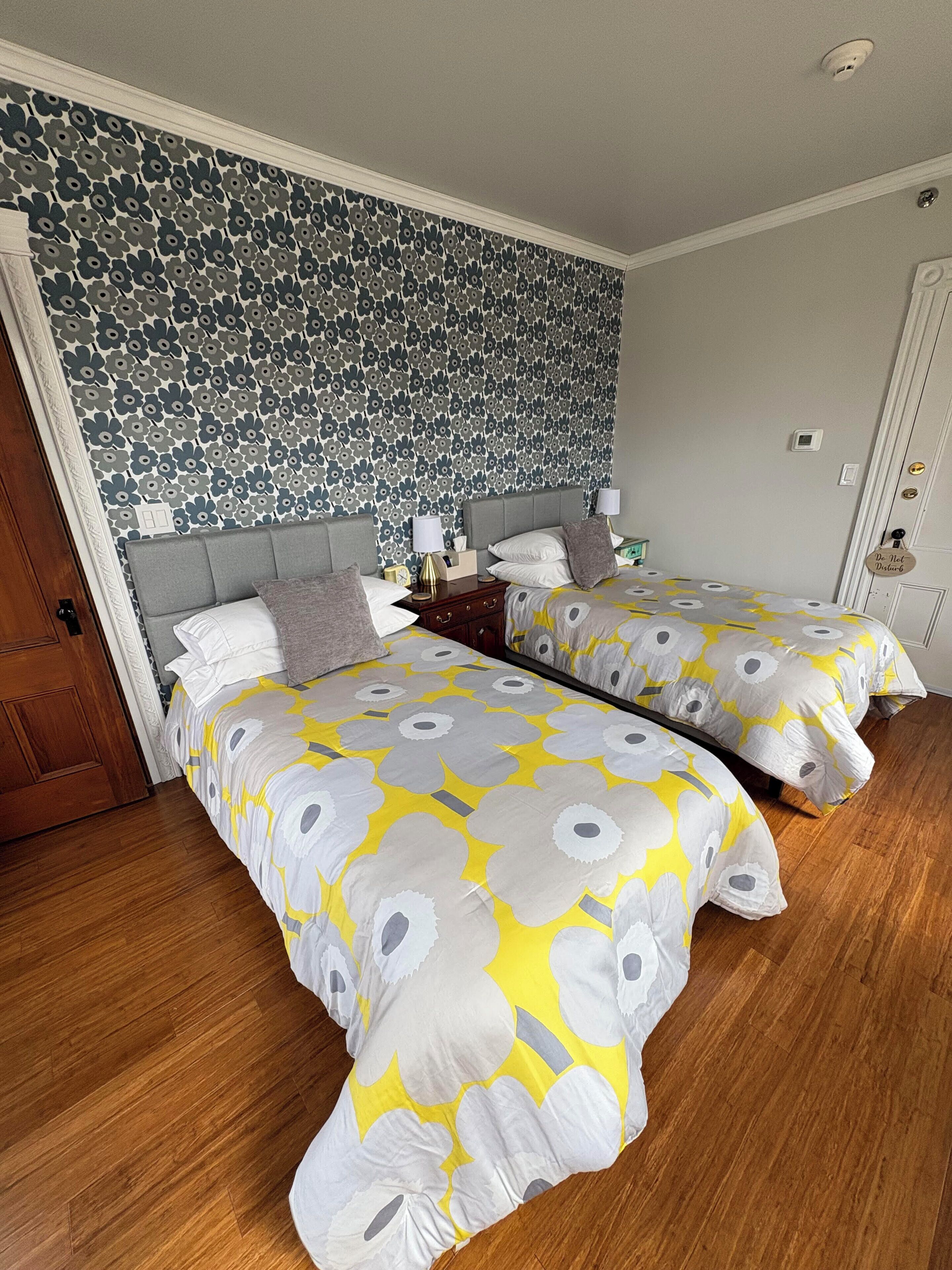A bedroom with two beds, a patterned wallpaper, a wooden floor, and a door with a doorknob.