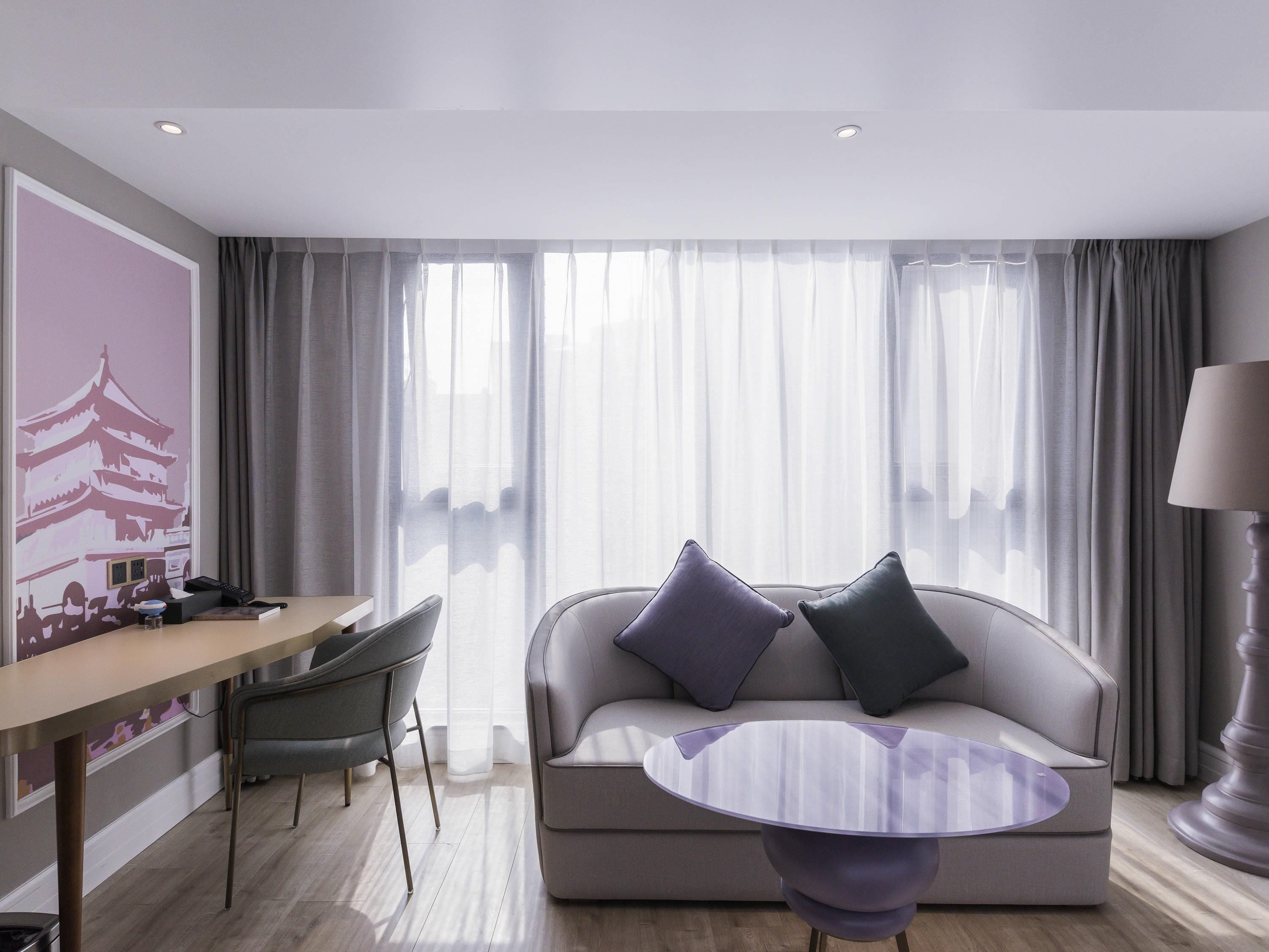 Una habitación de hotel moderna con sofá, una mesa redonda, un escritorio con silla y una lámpara.