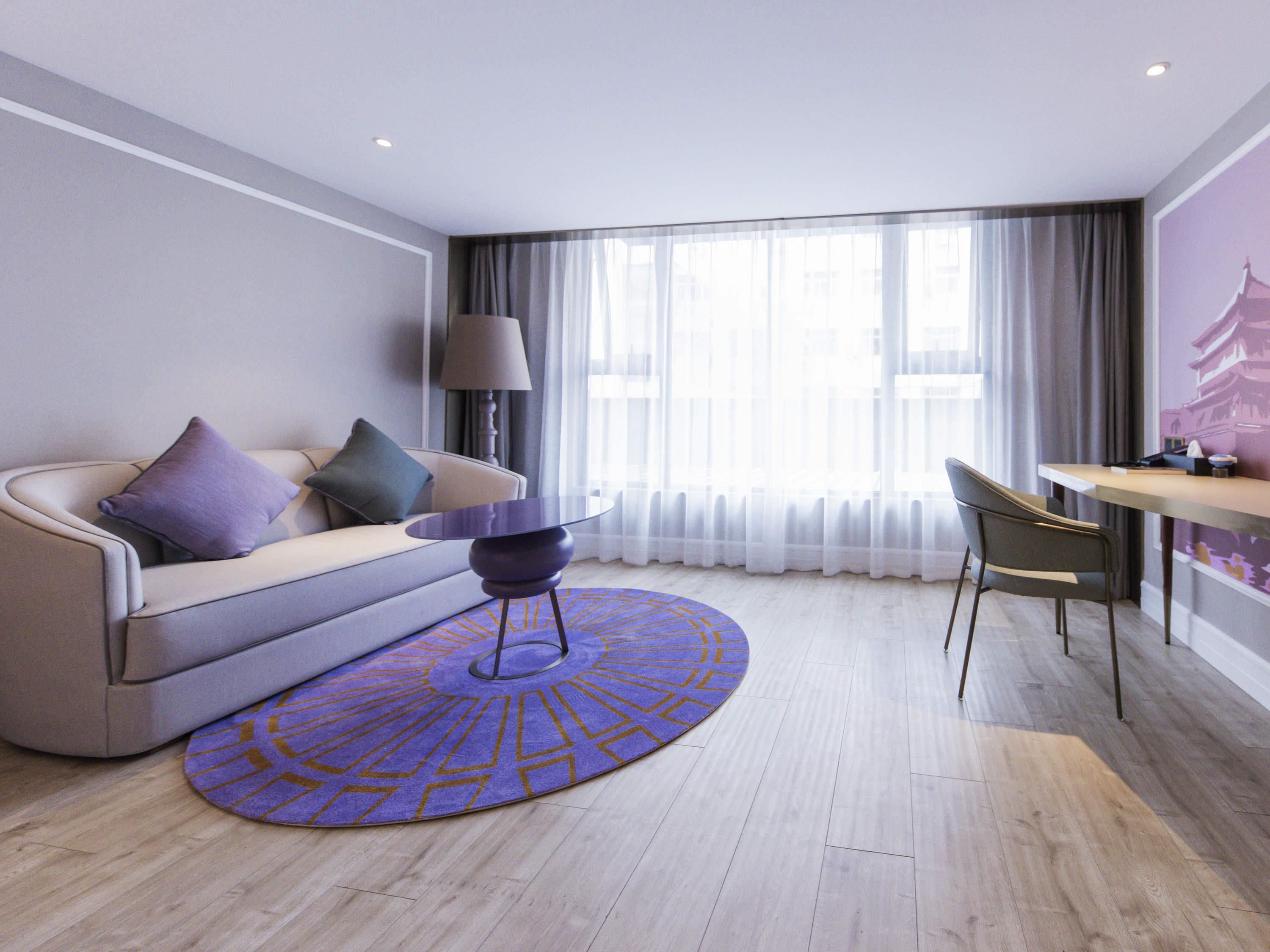 A modern hotel room with a sofa, a small round table, a chair, and a large window with curtains.