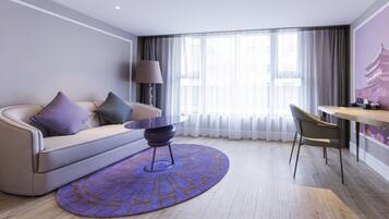 A modern hotel room with a sofa, a small round table, a chair, and a large window with curtains.
