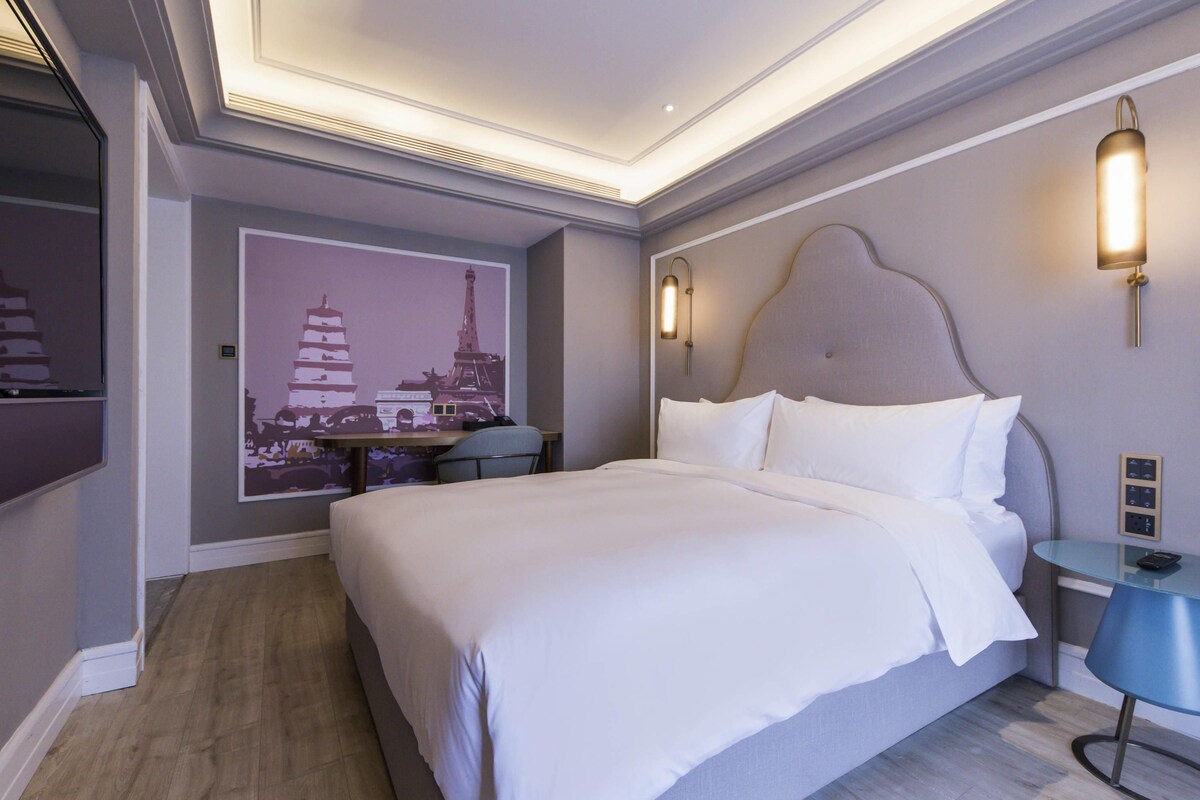 A modern hotel room with a large bed, a desk, a chair, and a wall-mounted TV.