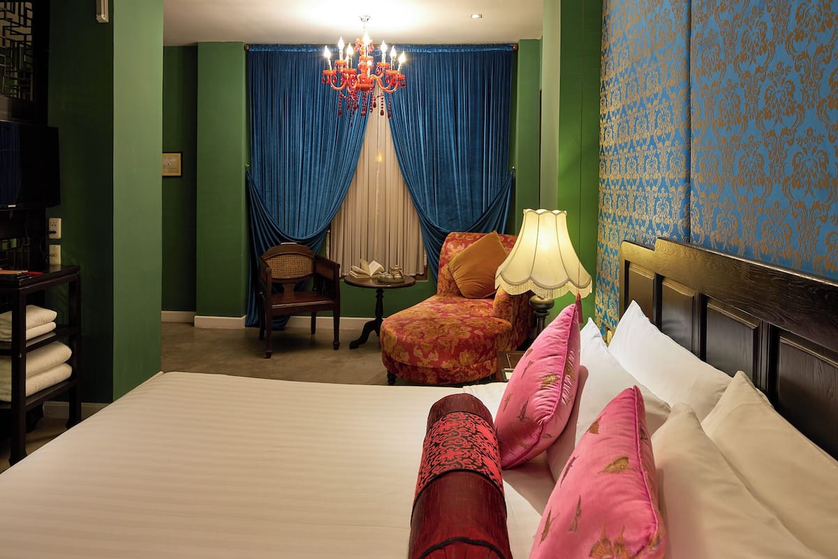 Quarto de hotel com uma cama grande, uma cadeira, uma mesa pequena e um candelabro.