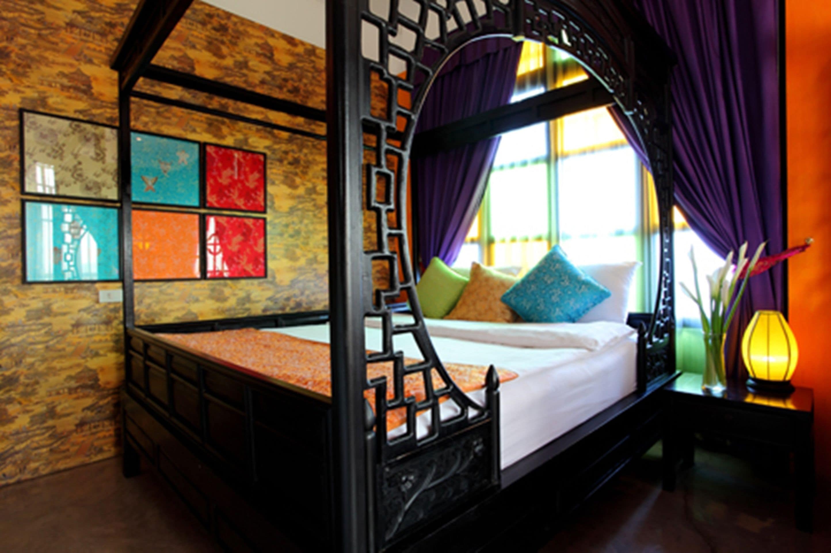 A four-poster bed with a wooden frame, a white mattress, and colorful pillows. There is a window with purple curtains and a vase with flowers on a nightstand beside the bed.