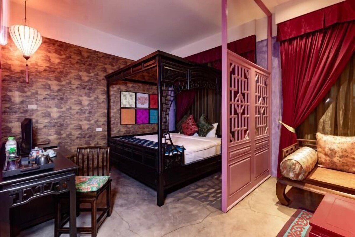 A four-poster bed with a wooden frame, a desk with a chair, a sofa, and a decorative wall with a mix of patterns.