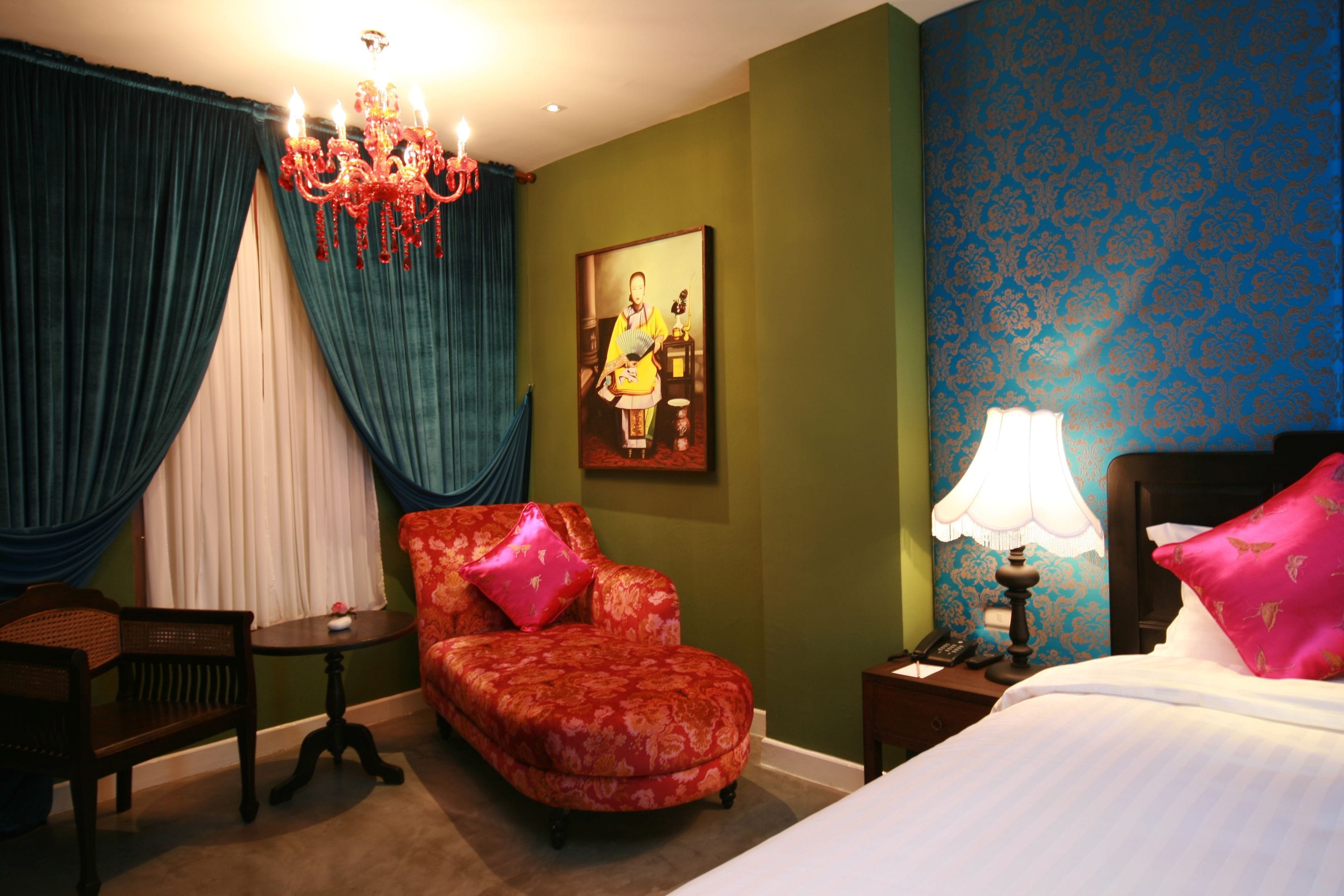 A hotel room with a bed, a chair, a table, a lamp, and a painting on the wall.